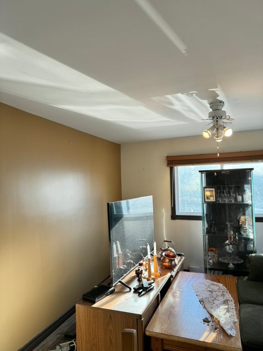 Salon avec murs beiges, télévision, ventilateur de plafond, armoire, fenêtre et table en bois.