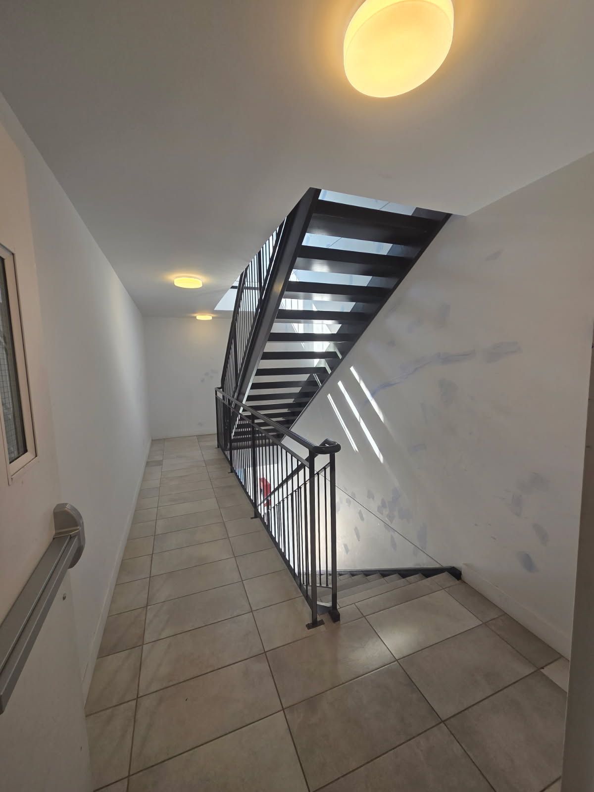 Escalier dans un couloir blanc avec une rampe en métal. La lumière du soleil inonde les marches.