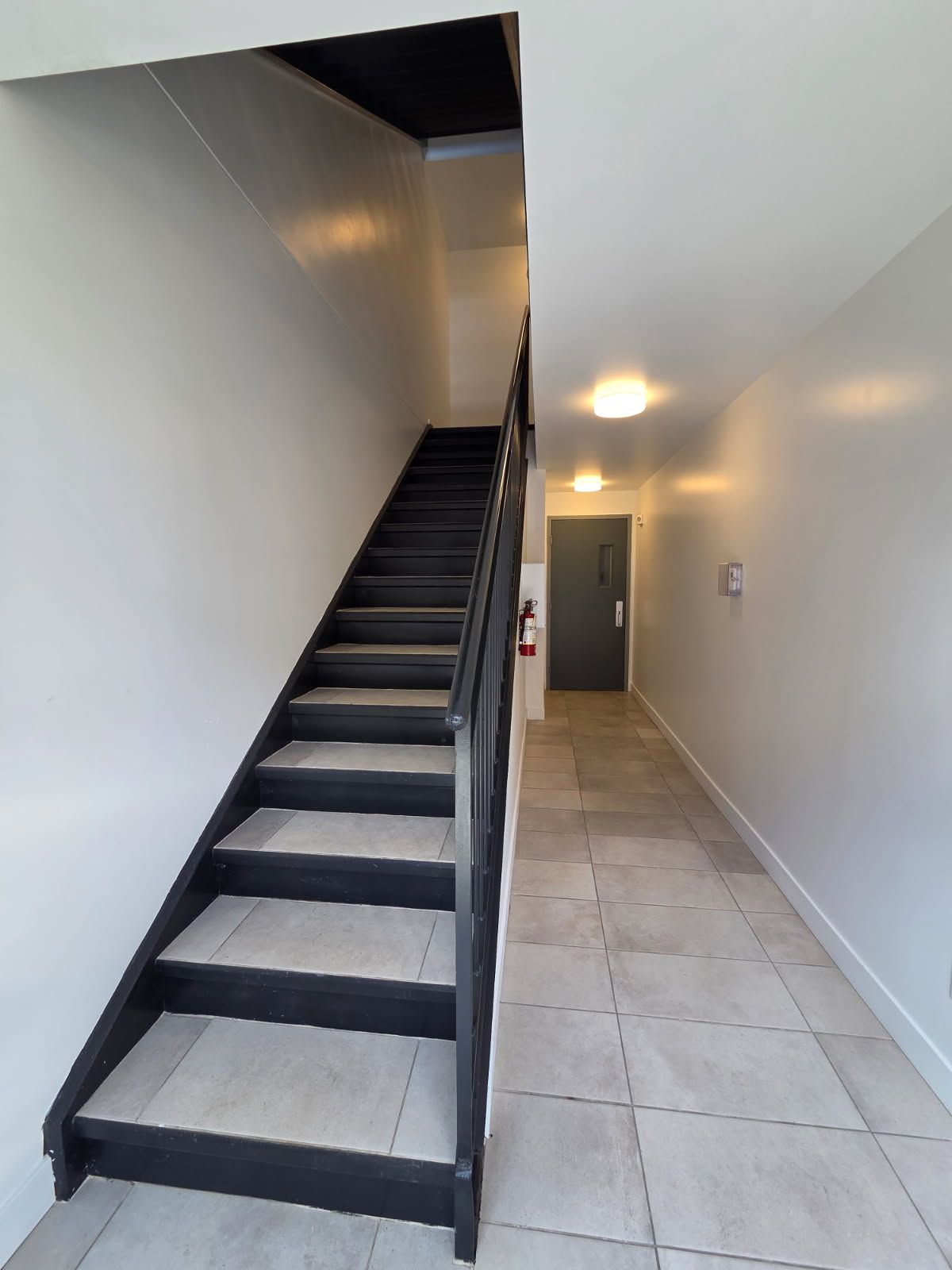 Escalier avec marches et rampe noires, menant à un couloir avec une porte. Sol carrelé gris.