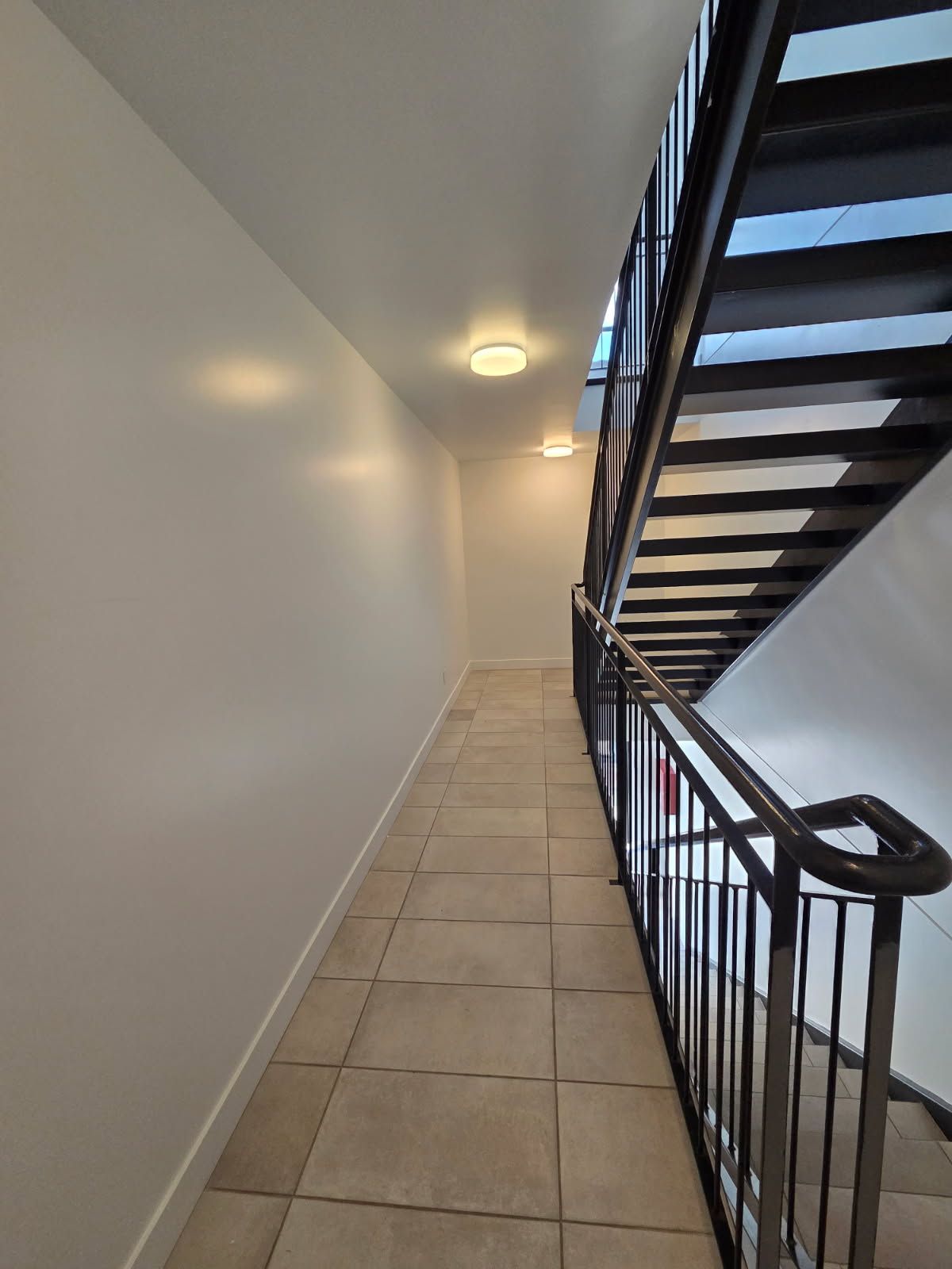 Cage d'escalier avec sol carrelé, murs blancs, rampe noire et éclairage au plafond.