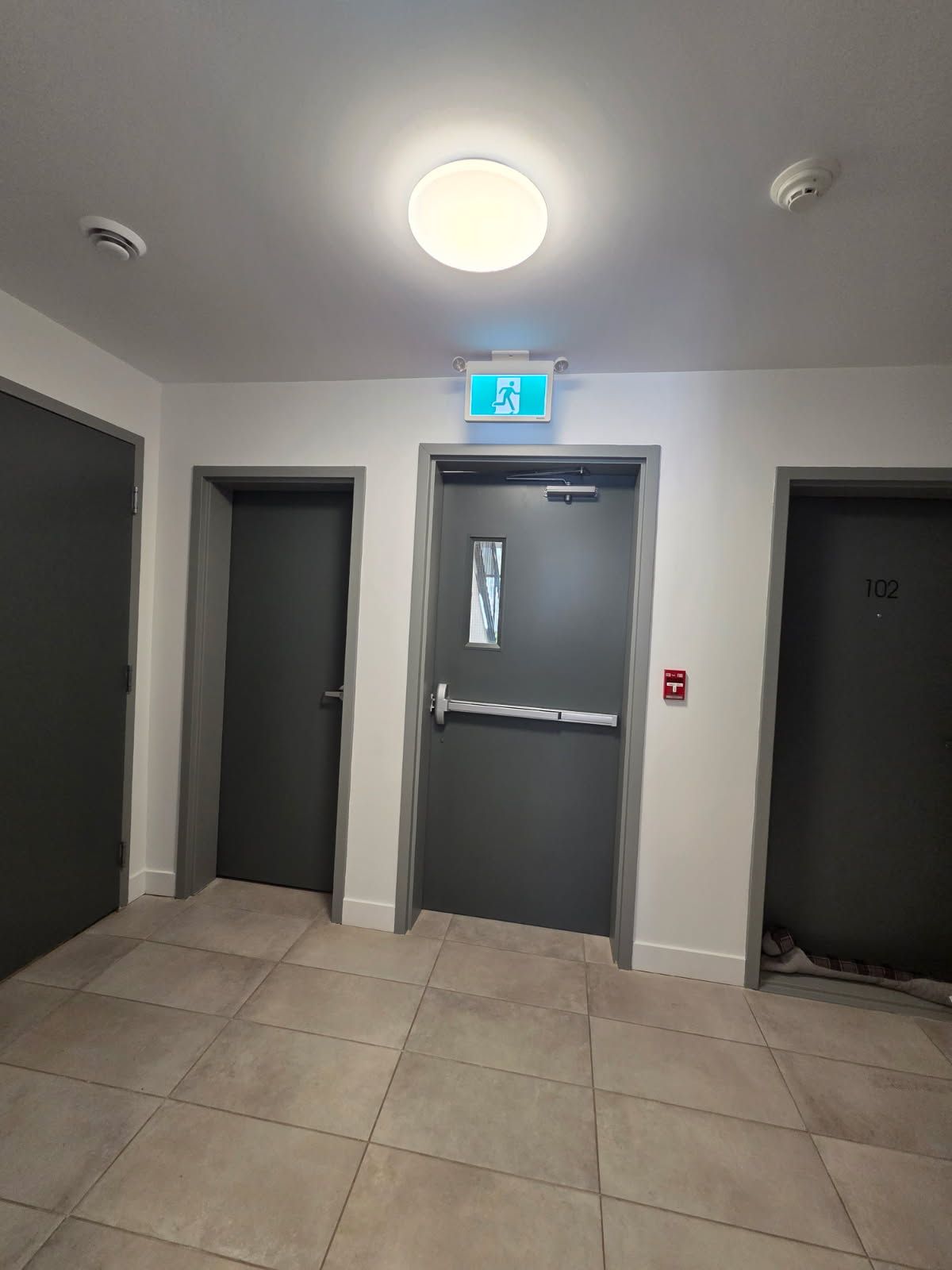 Couloir avec trois portes grises, panneau de sortie au-dessus d'une porte centrale et luminaire au plafond.
