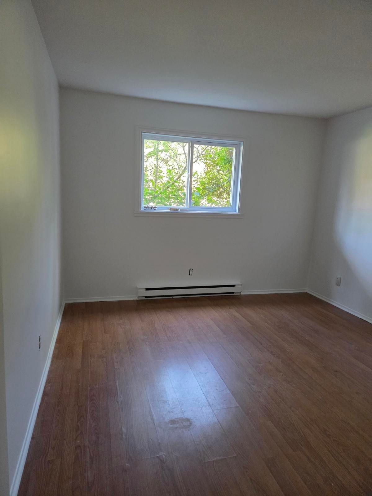 Pièce vide avec un sol imitation bois, une fenêtre et un appareil de chauffage en dessous.