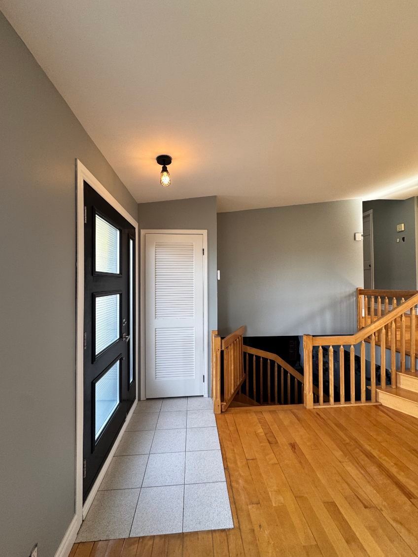 Entrée avec murs gris, parquet, porte et escalier. Fenêtre à cadre noir et petit plafonnier.