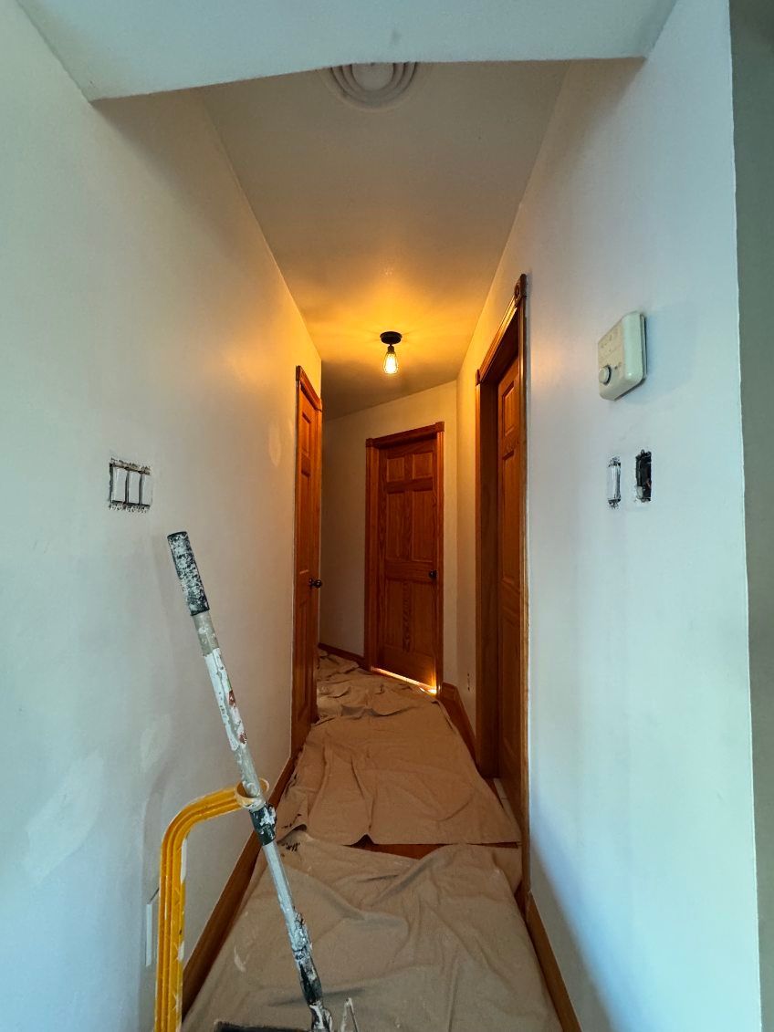 Couloir aux murs blancs fraîchement peints, portes marron et bâches de protection. Une échelle est appuyée contre le mur.