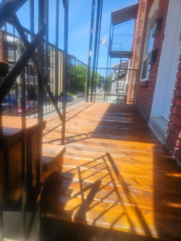 Terrasse en bois teintée en brun, rambarde noire et bâtiment en briques rouges baigné de soleil.
