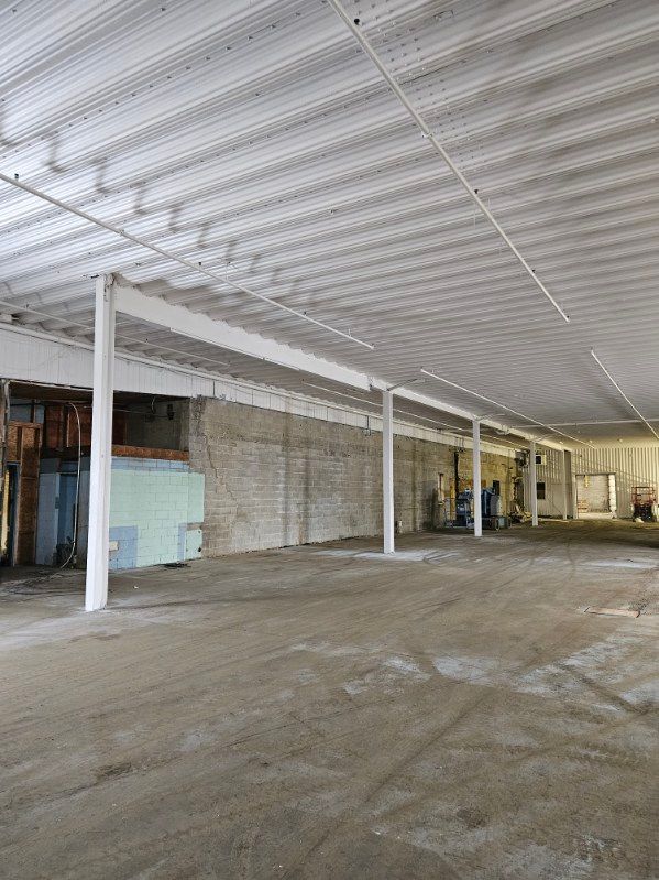 Espace industriel vide avec sol en béton, poutres blanches et plafond en tôle ondulée.