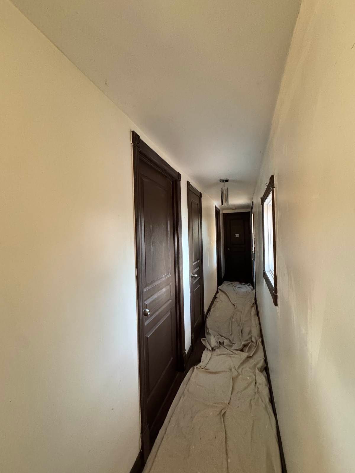 Couloir étroit aux murs beiges, portes marron et sol recouvert.
