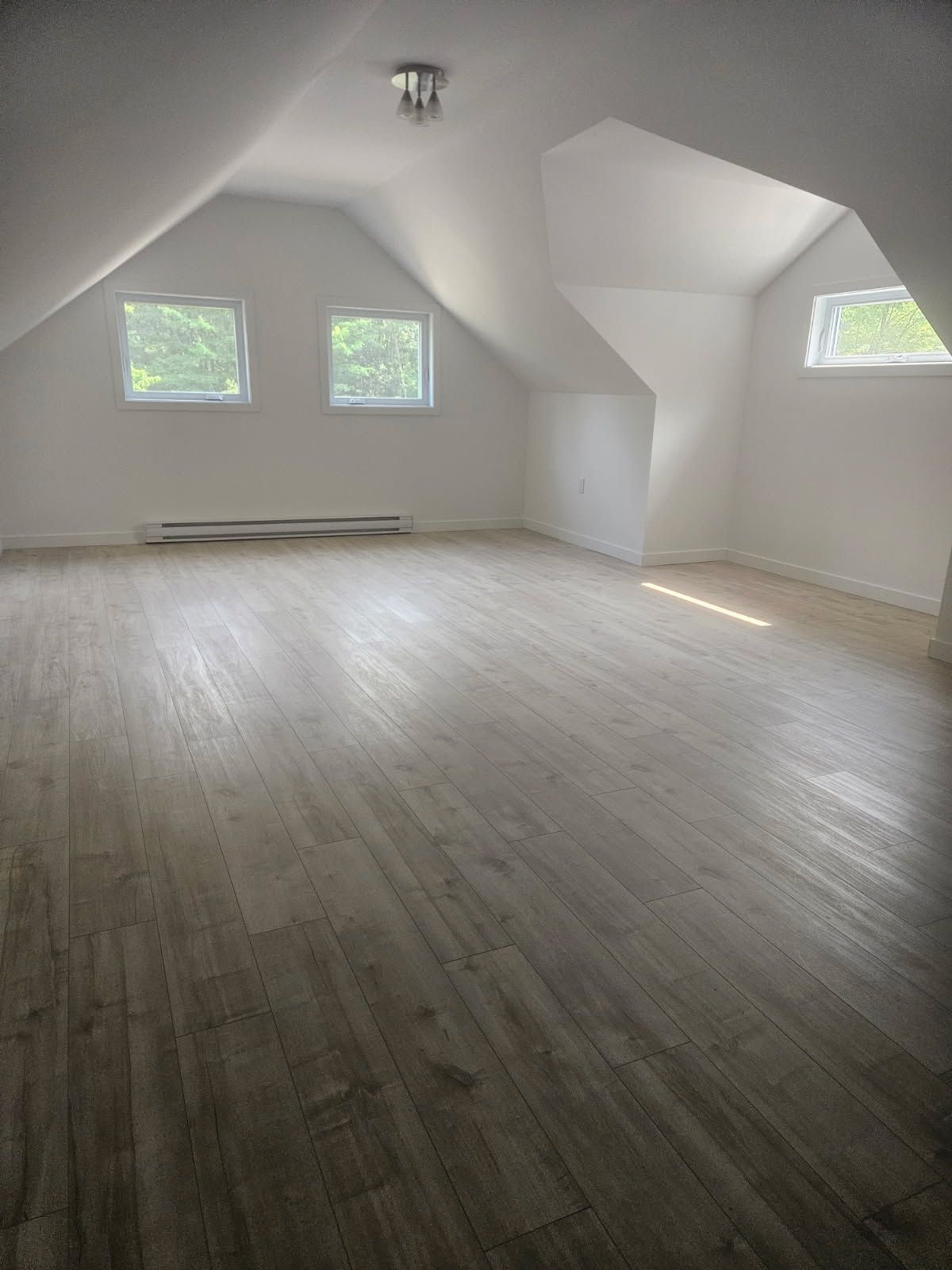 Pièce mansardée vide avec un sol imitation bois, des murs blancs en pente, de petites fenêtres et un plafonnier.