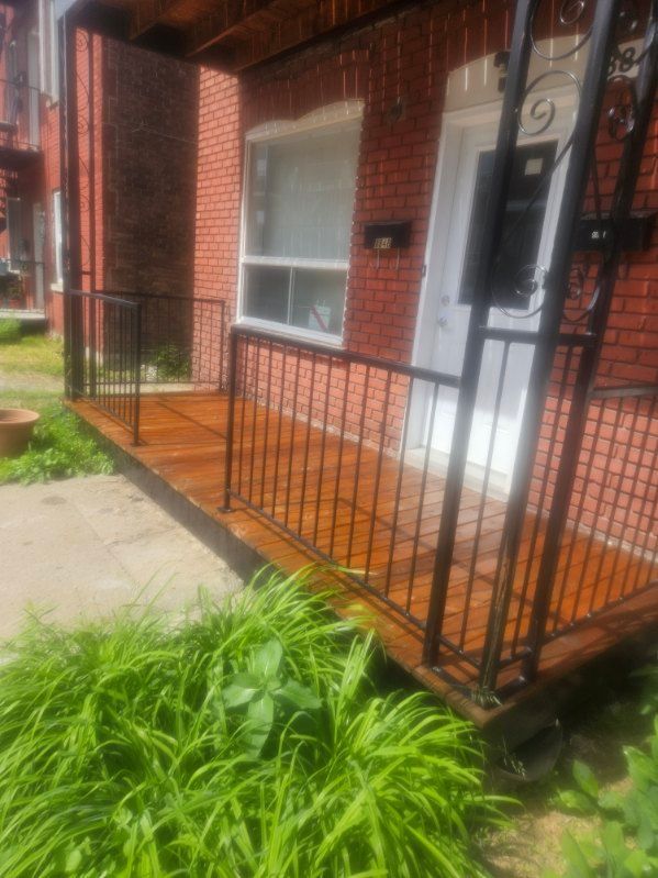 Porche en bois avec balustrade en métal noir devant un bâtiment en briques rouges.
