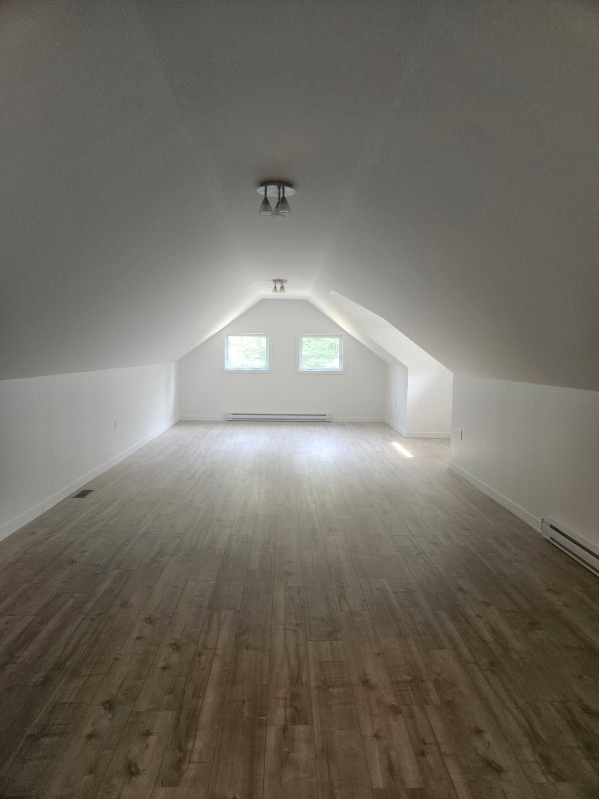 Pièce mansardée vide avec murs blancs, plafond mansardé, deux fenêtres et sol imitation bois.