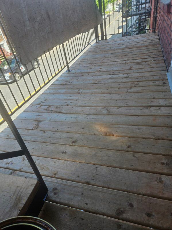 Terrasse en bois avec rambarde métallique, adossée à un mur de briques. La lumière du soleil illumine les planches.