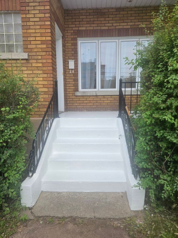 Maison en briques avec marches en béton peint en blanc et rambarde noire. Des buissons verts encadrent l'entrée.