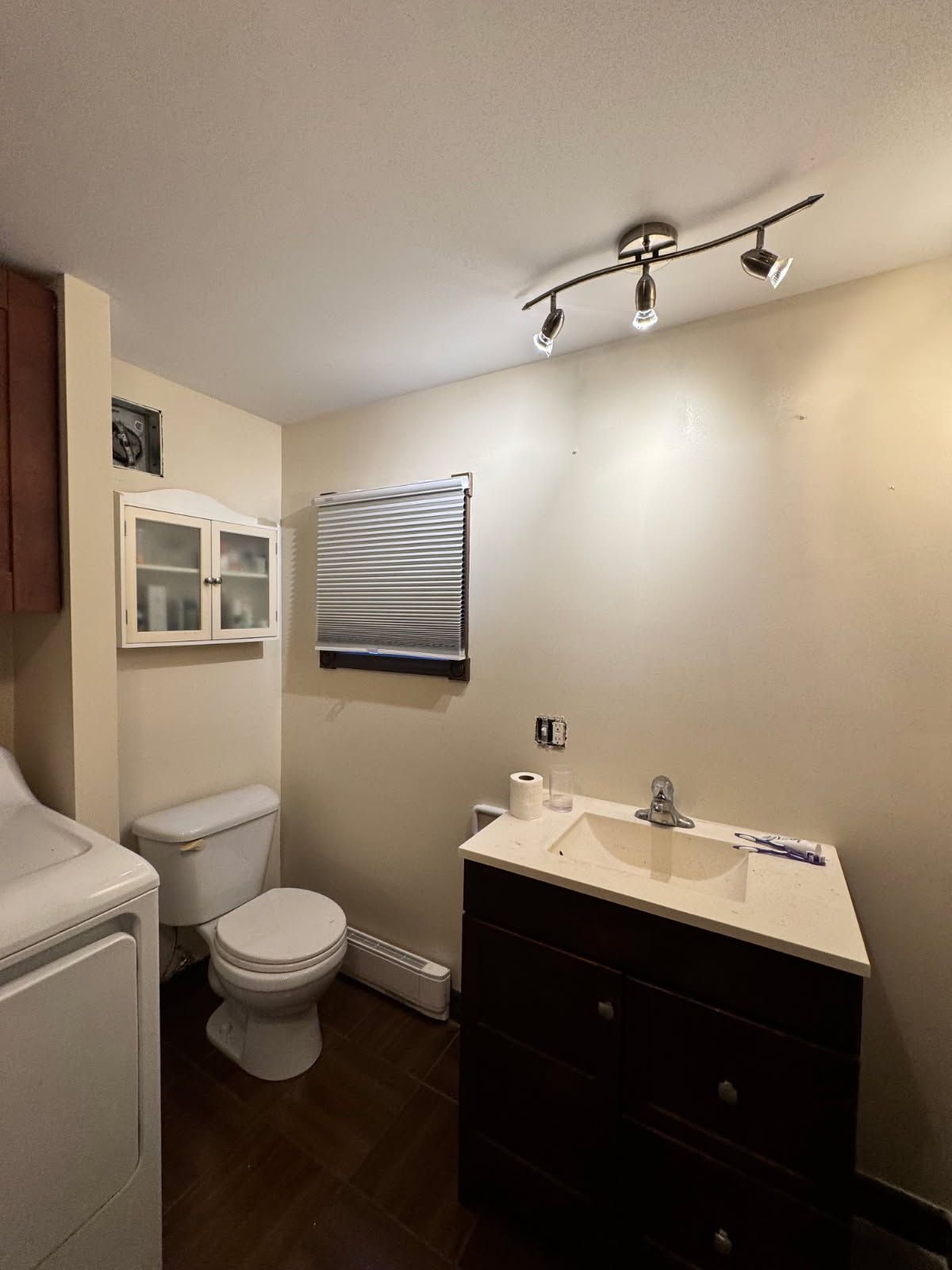 Salle de bains avec toilettes, meuble-lavabo et armoire ; murs neutres, sol foncé et éclairage au plafond.