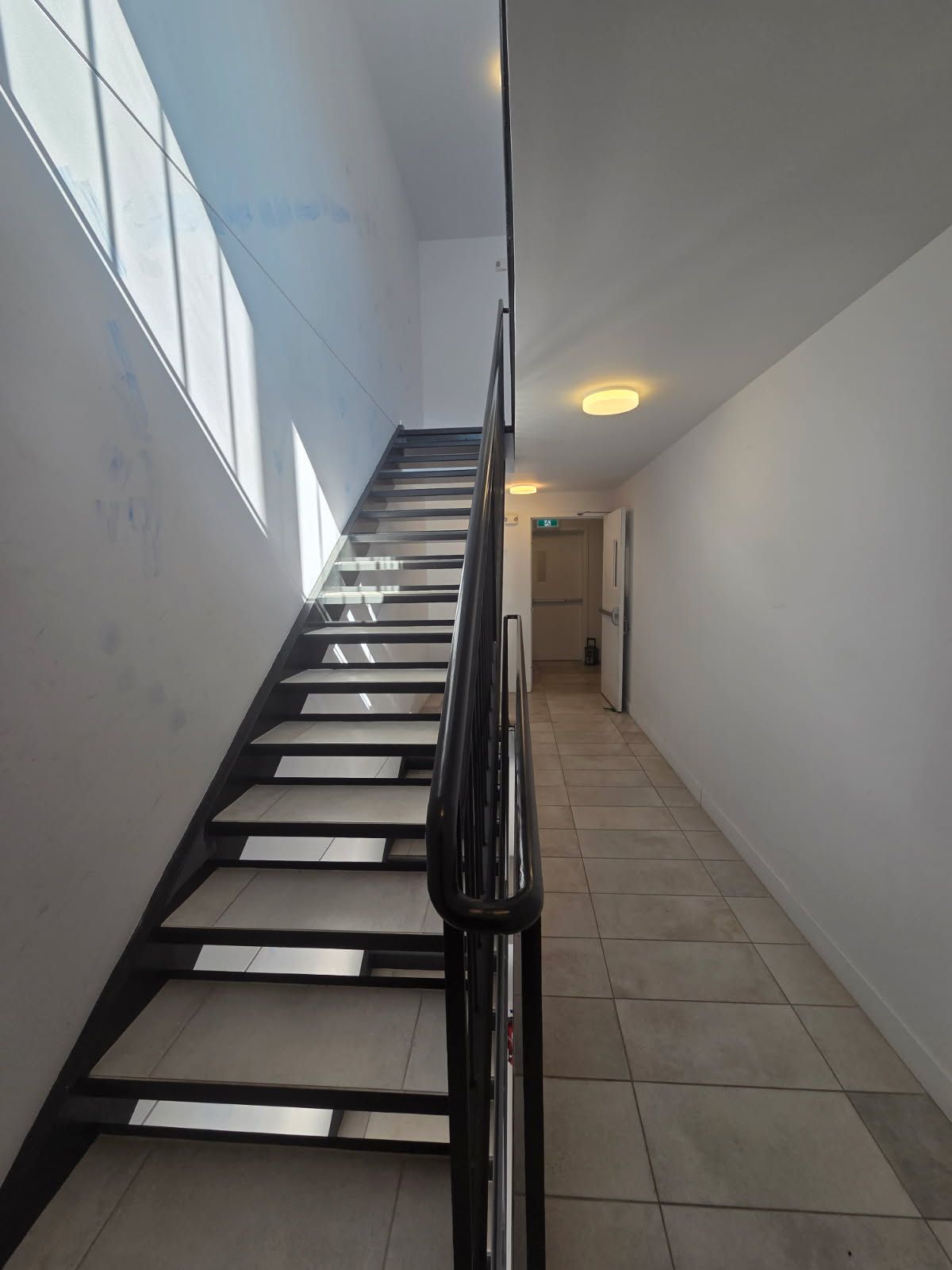 Un escalier avec une rampe menant à l'étage. La lumière inonde l'espace par les fenêtres. À droite, un couloir carrelé.