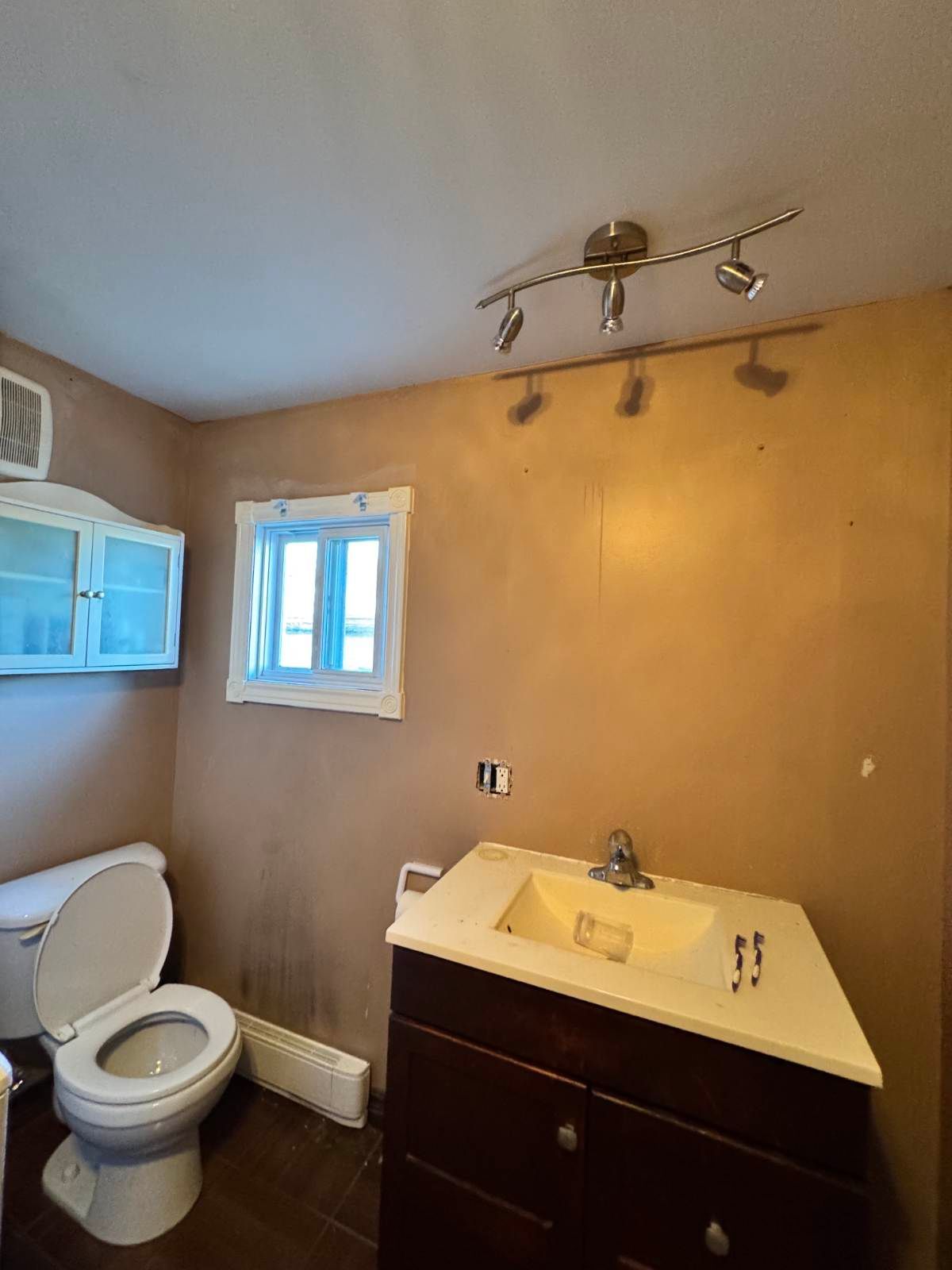 Salle de bains avec toilettes, meuble-lavabo et fenêtre ; murs beiges et spot lumineux sur rail.