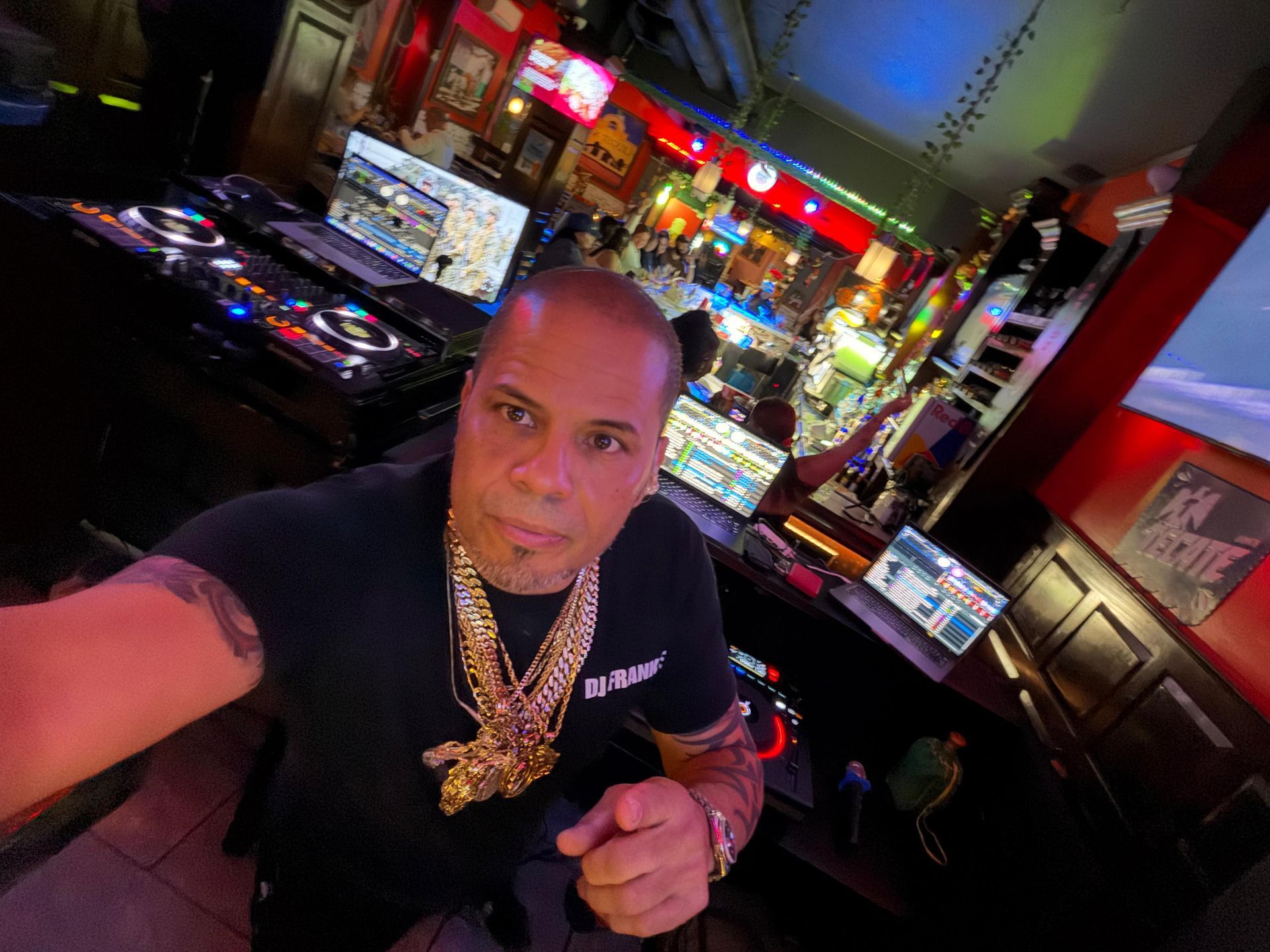Man taking a selfie in a brightly lit bar, wearing gold chains and leaning toward the camera.