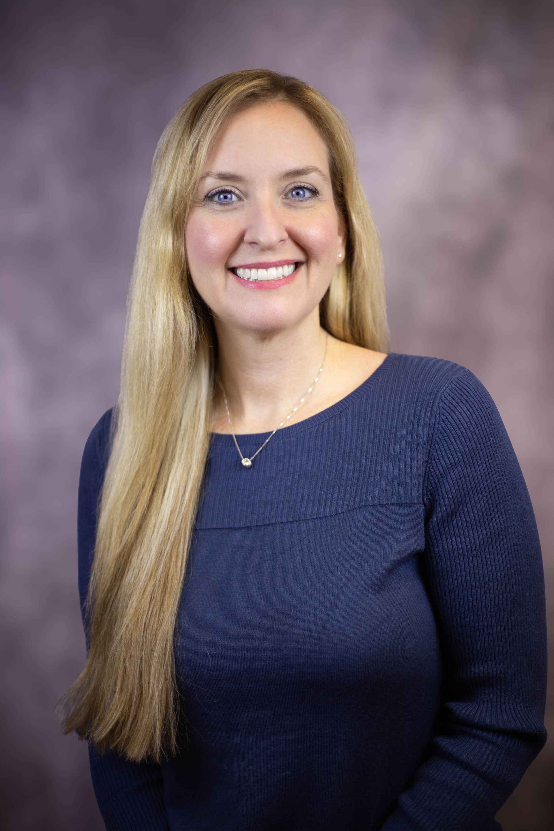 A woman wearing a blue sweater and a necklace is smiling for the camera.
