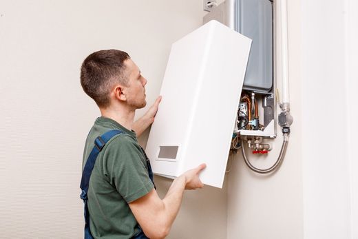 Man installing a white boiler unit on a wall. He's wearing work clothes, focused on the task.