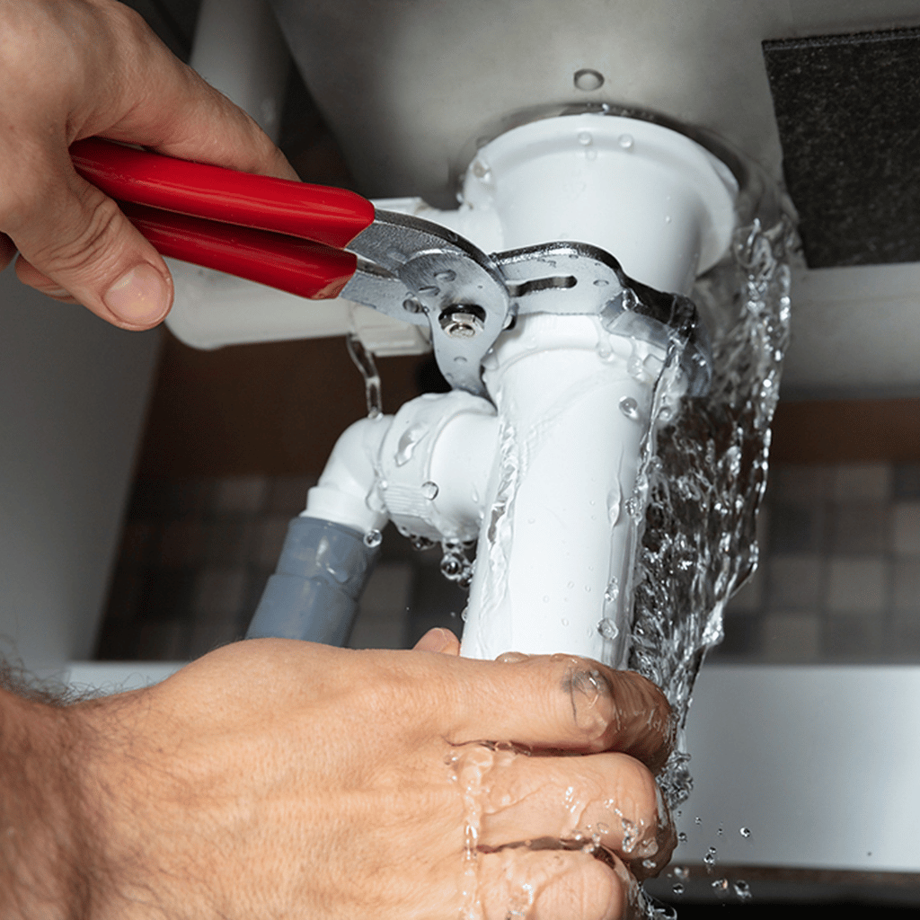 Hands using pliers to tighten a leaking white plastic sink drainpipe under a sink. Water streams out.