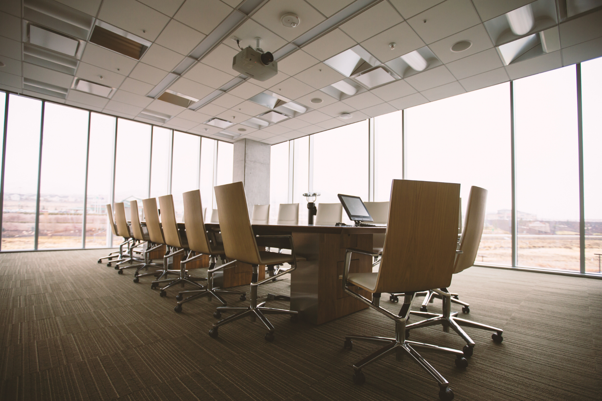 A modern, empty boardroom with a long wooden table surrounded by tan chairs, featuring floor-to-ceiling windows.