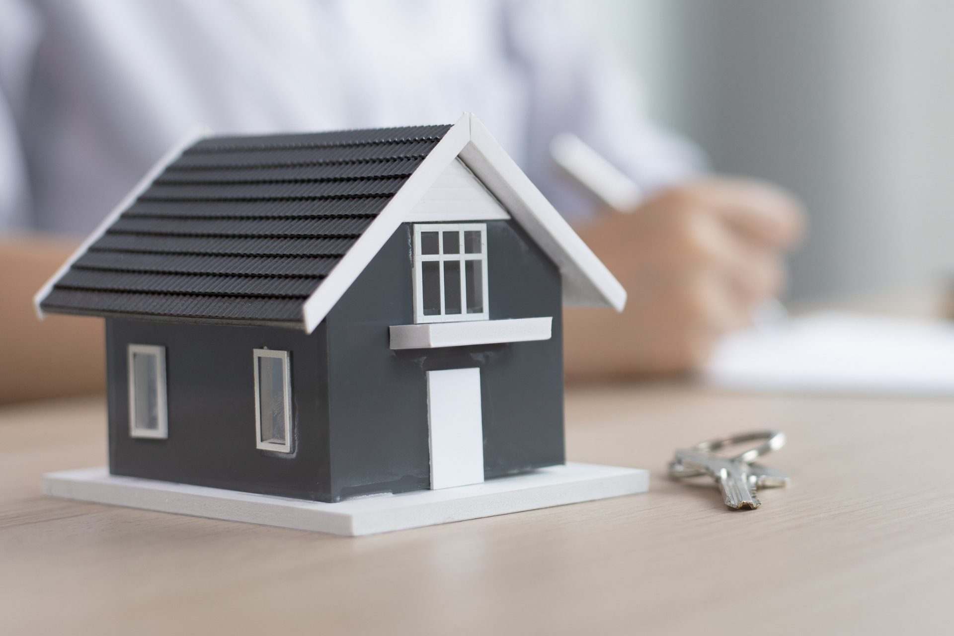 A small model house sitting on a desk next to a set of keys, with a person writing in the background.
