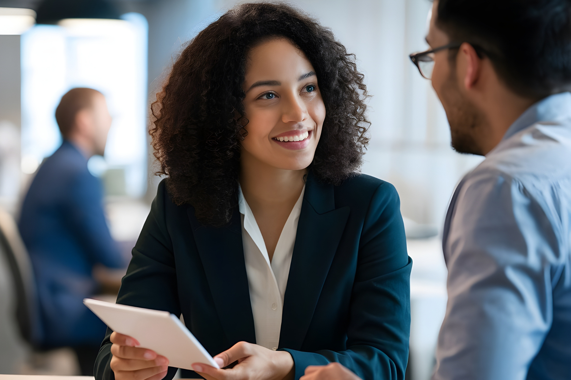 A smiling professional holds a tablet and speaks to a colleague in an office setting.