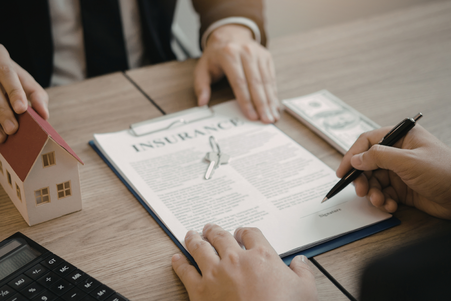 Two people sign an insurance document on a desk featuring a small house model, keys, cash, and a calculator.