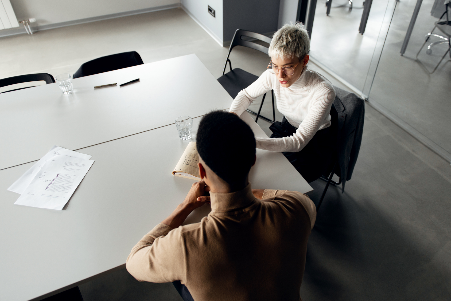 Two people in neutral clothing sit at a table in a modern, brightly lit office, engaged in a conversation.