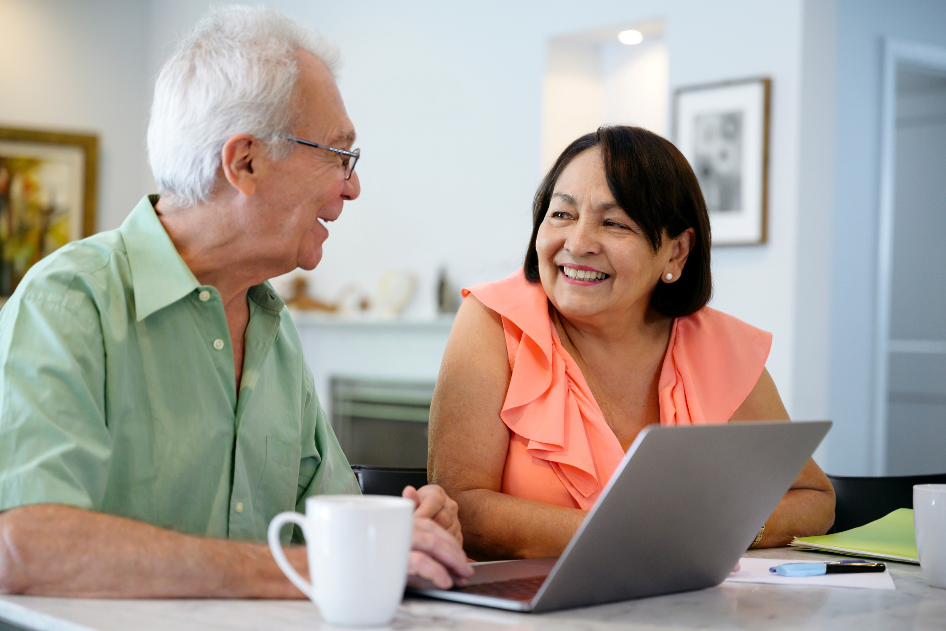 Two people looking at a laptop together at a table with coffee mugs in a brightly lit room.