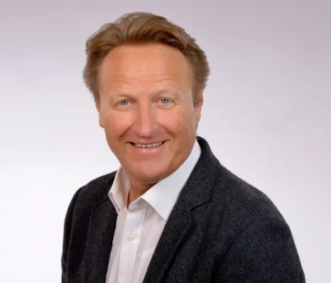 Ein Mann trägt ein weißes Hemd  mit Jackett und lächelt in die Kamera.