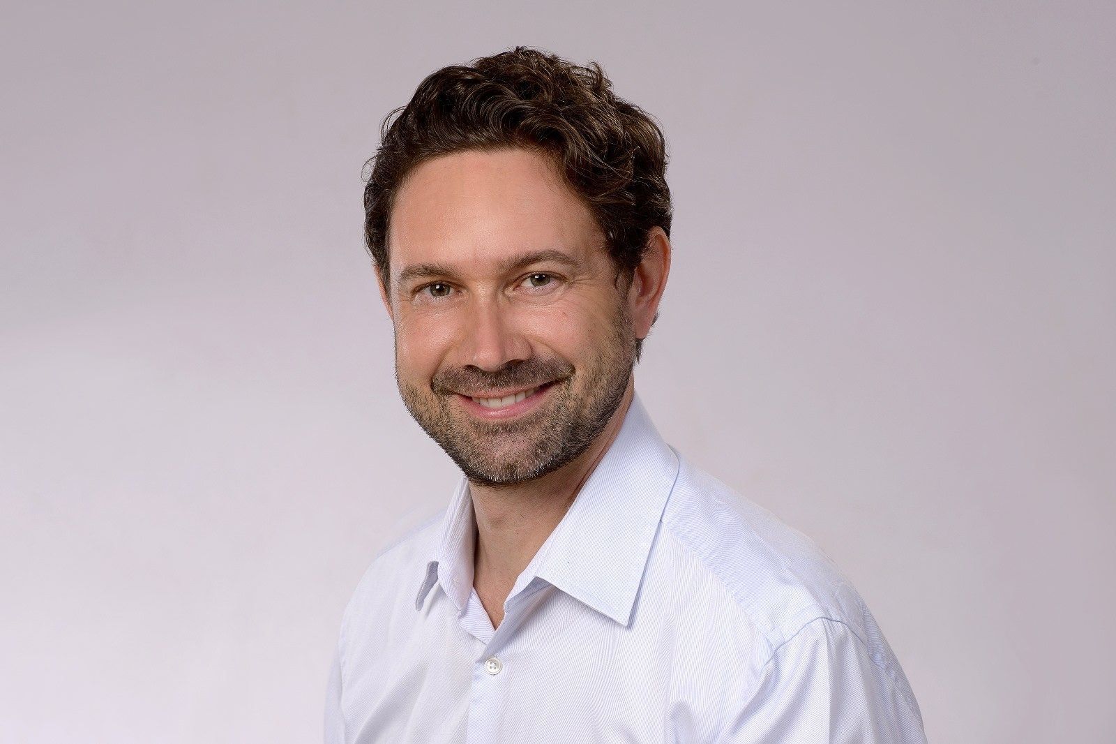 Ein Mann mit Bart trägt ein weißes Hemd und lächelt in die Kamera.