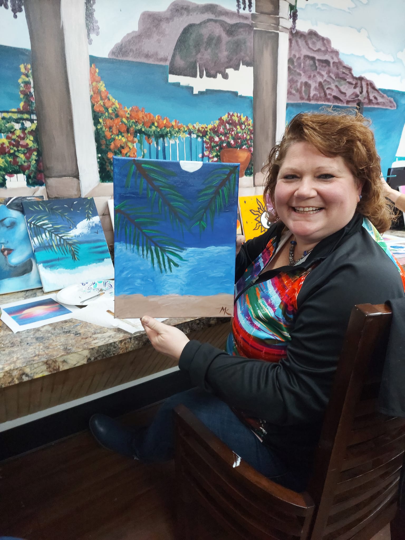 A woman is sitting in a chair holding a painting of a beach.