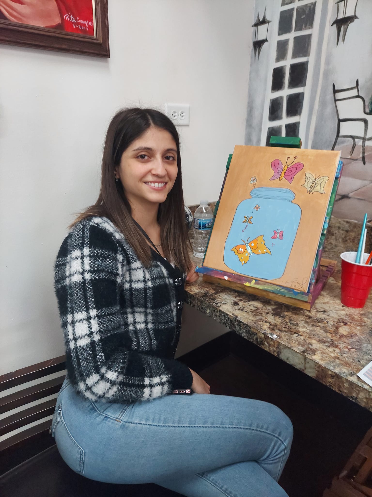 A woman is sitting in front of a painting on an easel.