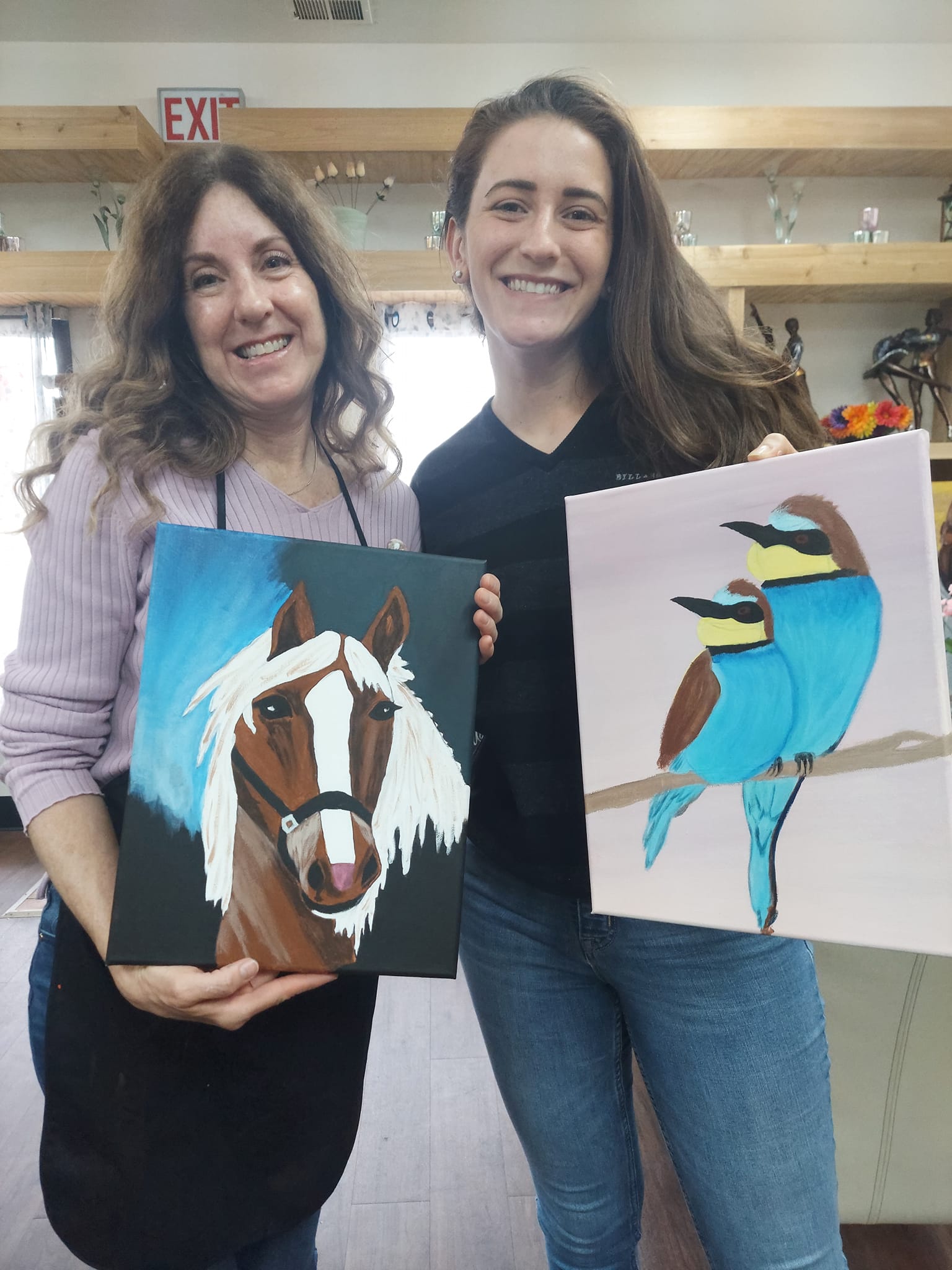 Two women are holding paintings of a horse and a bird.