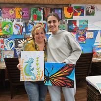 Two women are standing next to each other holding paintings of a seahorse and a butterfly.
