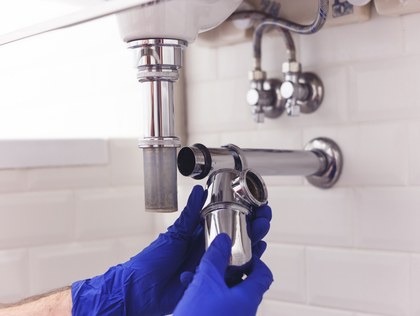 Gloved hands install a chrome faucet under a sink in a white-tiled bathroom.