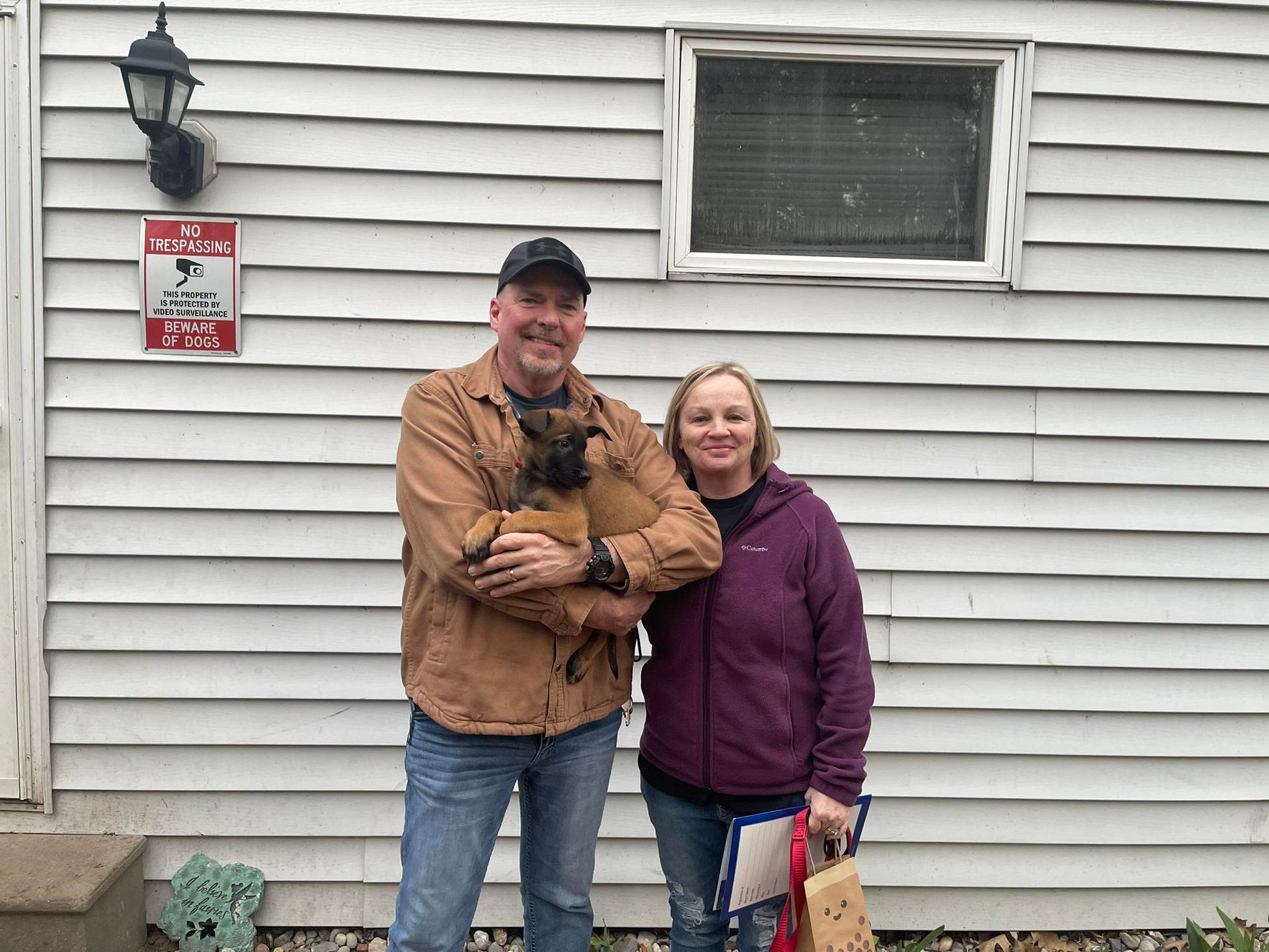 Man holding puppy, woman beside him in front of a white house. They smile.