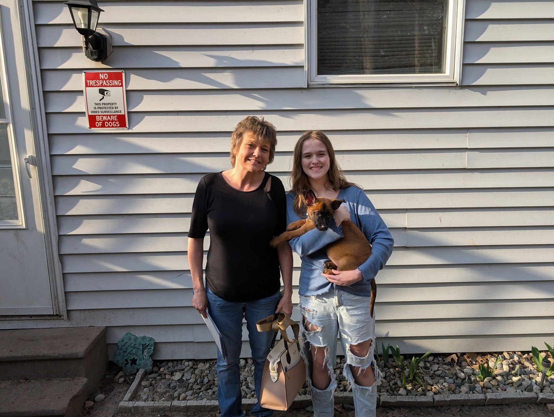 Two women with a puppy in front of a house. The woman on the right holds the puppy.