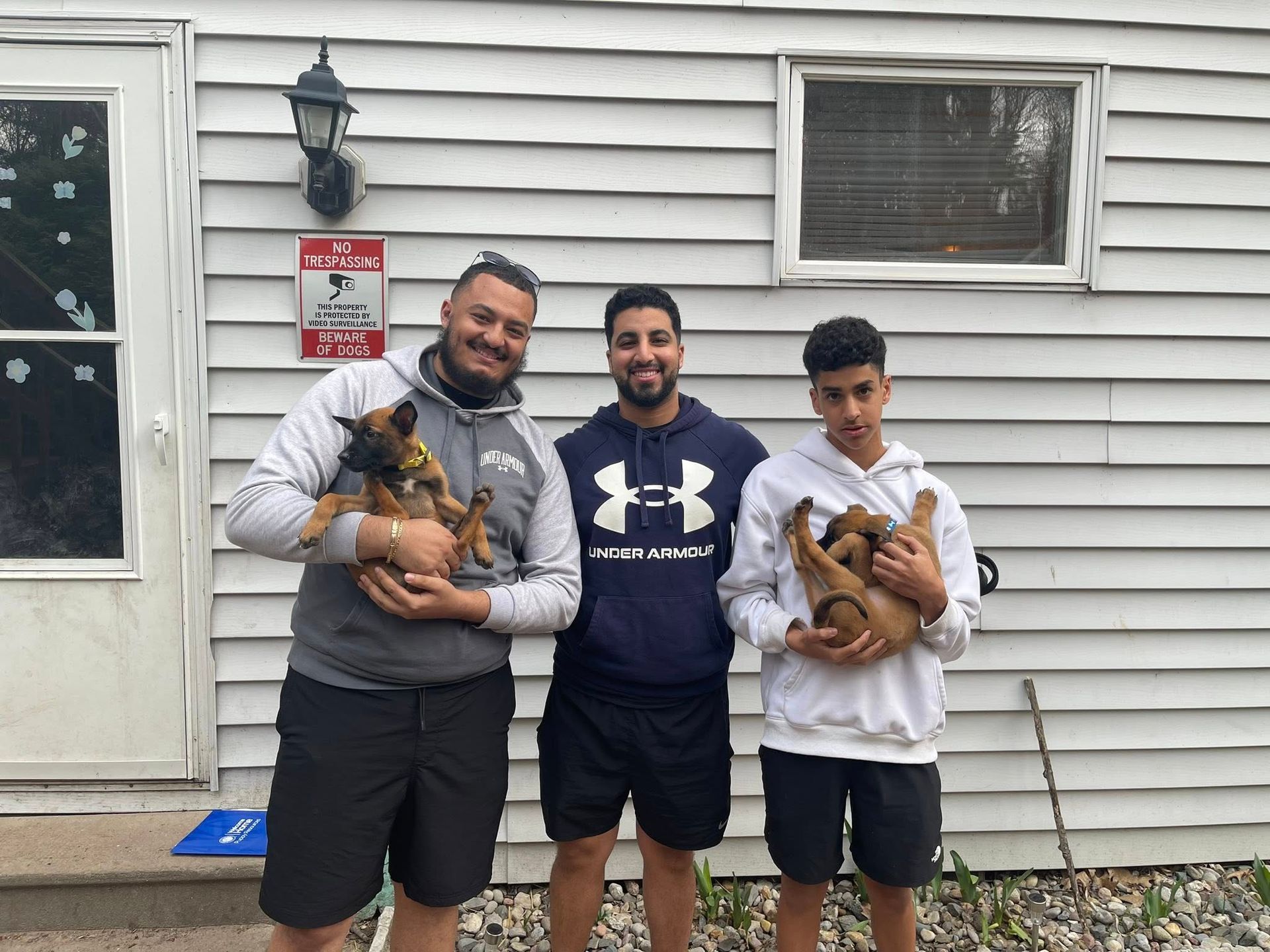 Three men outside holding puppies. One man smiles, all are near a house.