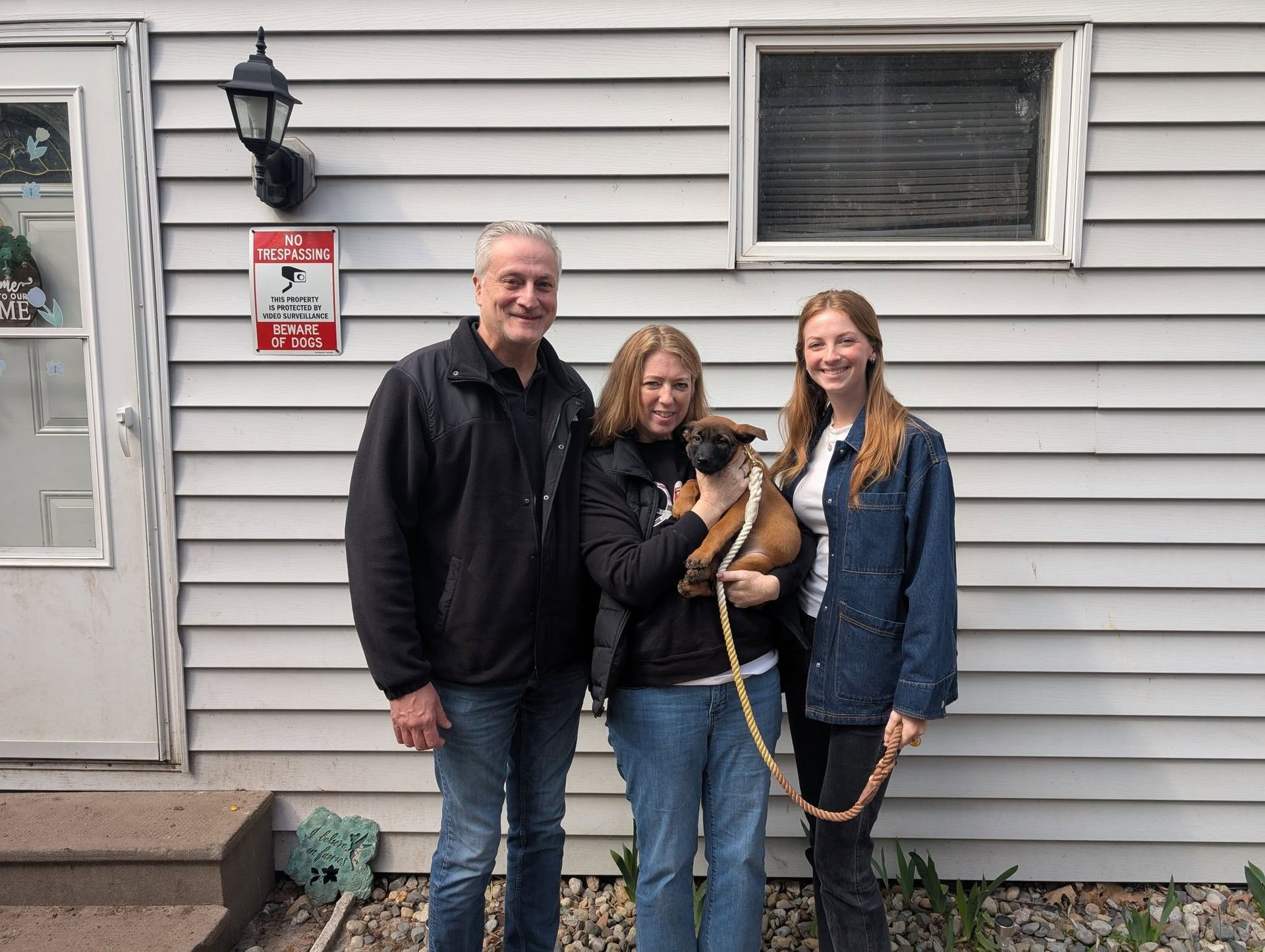A family, smiling, stands outside their house with a brown puppy.