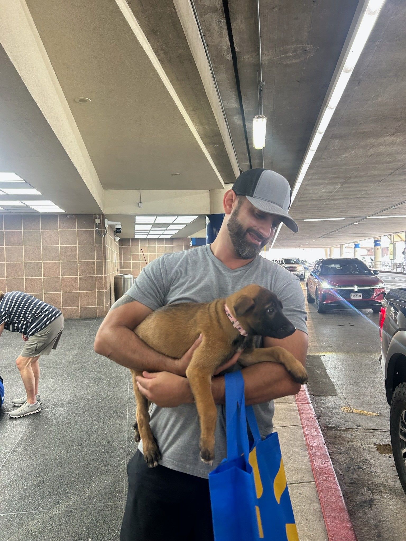 Man holding a brown puppy outside a store. He wears a hat and smiles. Another person is nearby.