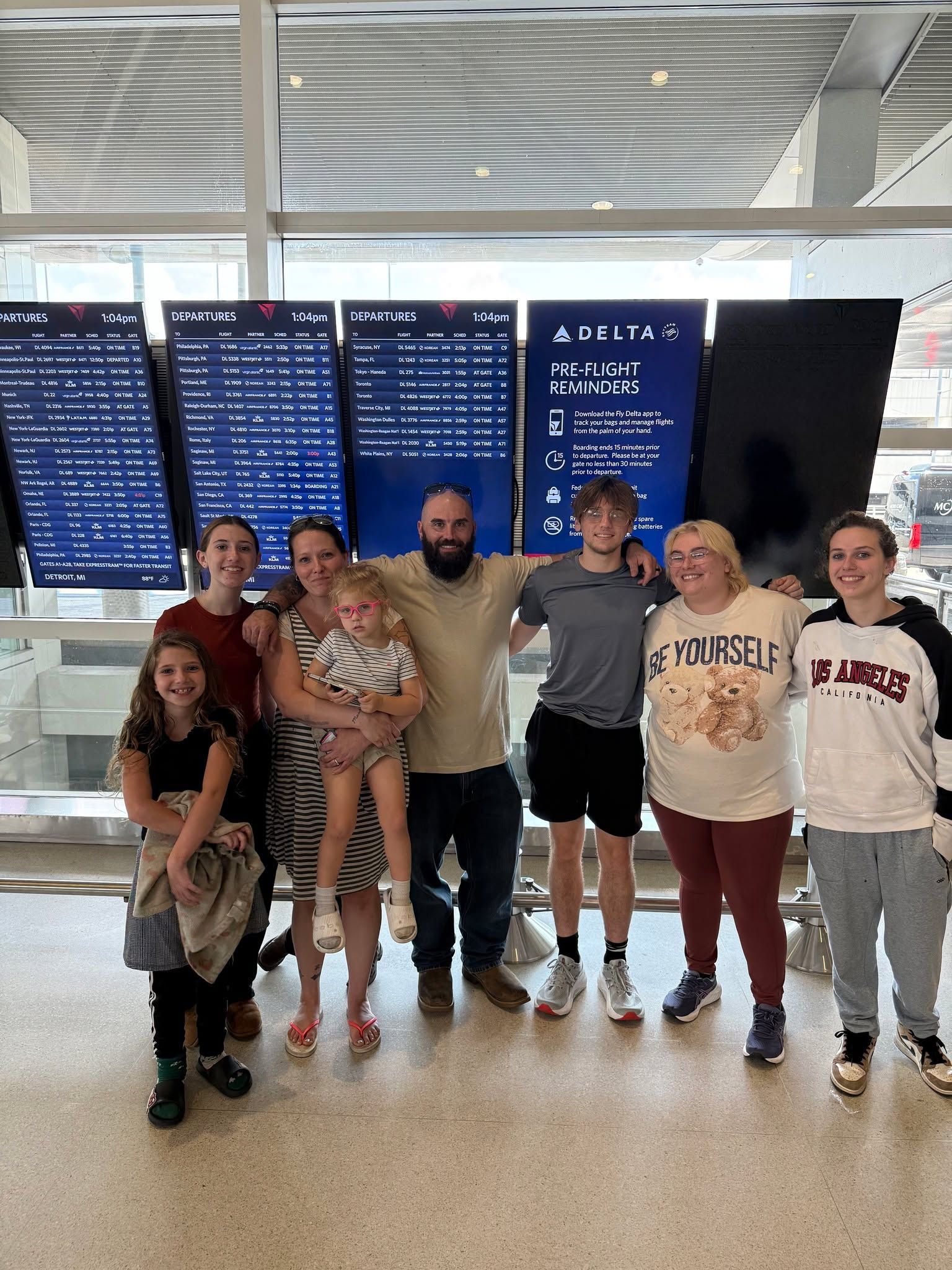 Family of eight poses in front of flight information screens at an airport. Smiling, arms around each other.