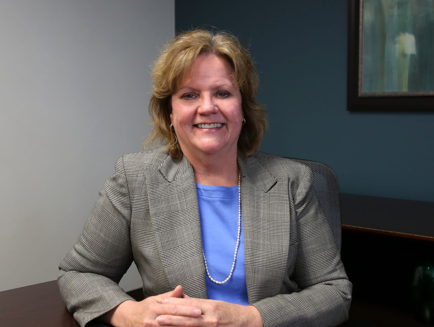 Woman in a business suit smiles at the camera, sitting at a desk in an office.