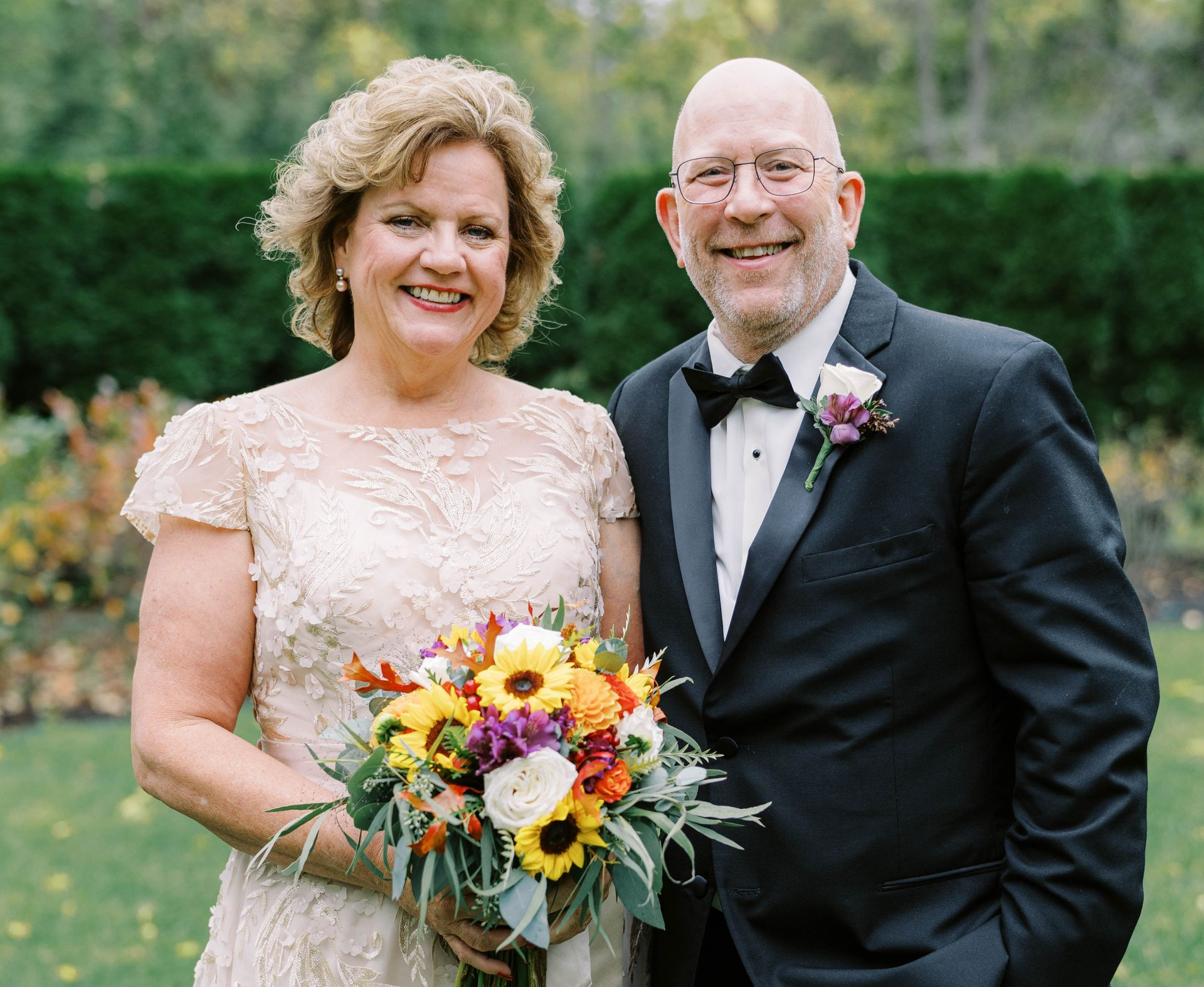 Couple smiling at camera; woman holds bouquet, man in tuxedo outdoors.