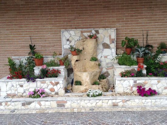 Dei muretti in pietra con dei vasi di fiori, delle piante interrate e al centro una pietra a forma di tronco con sopra della piantine
