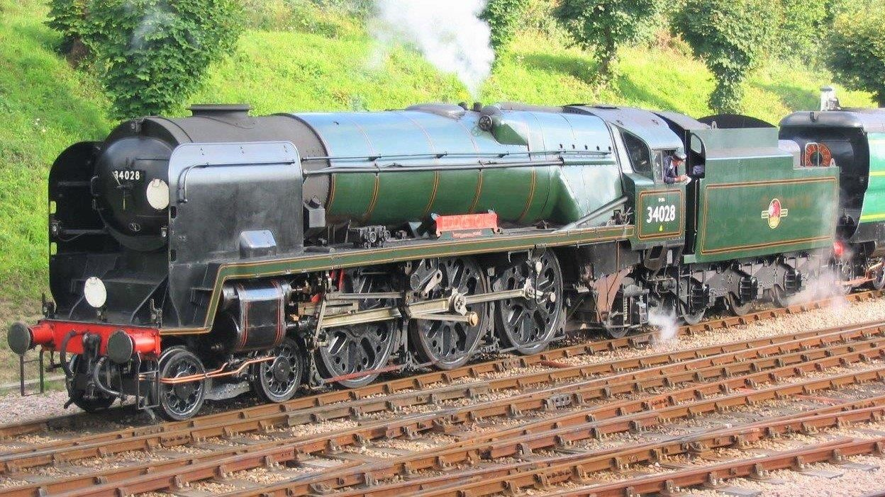 Steam train traveling alongside a canal, emitting smoke against a blue sky.