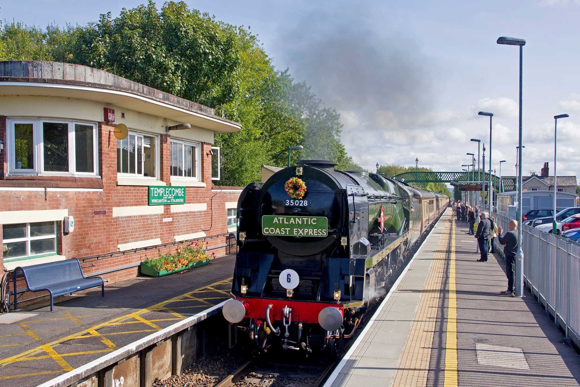 Steam train at station platform, with brick building and green sign. Passengers watch.