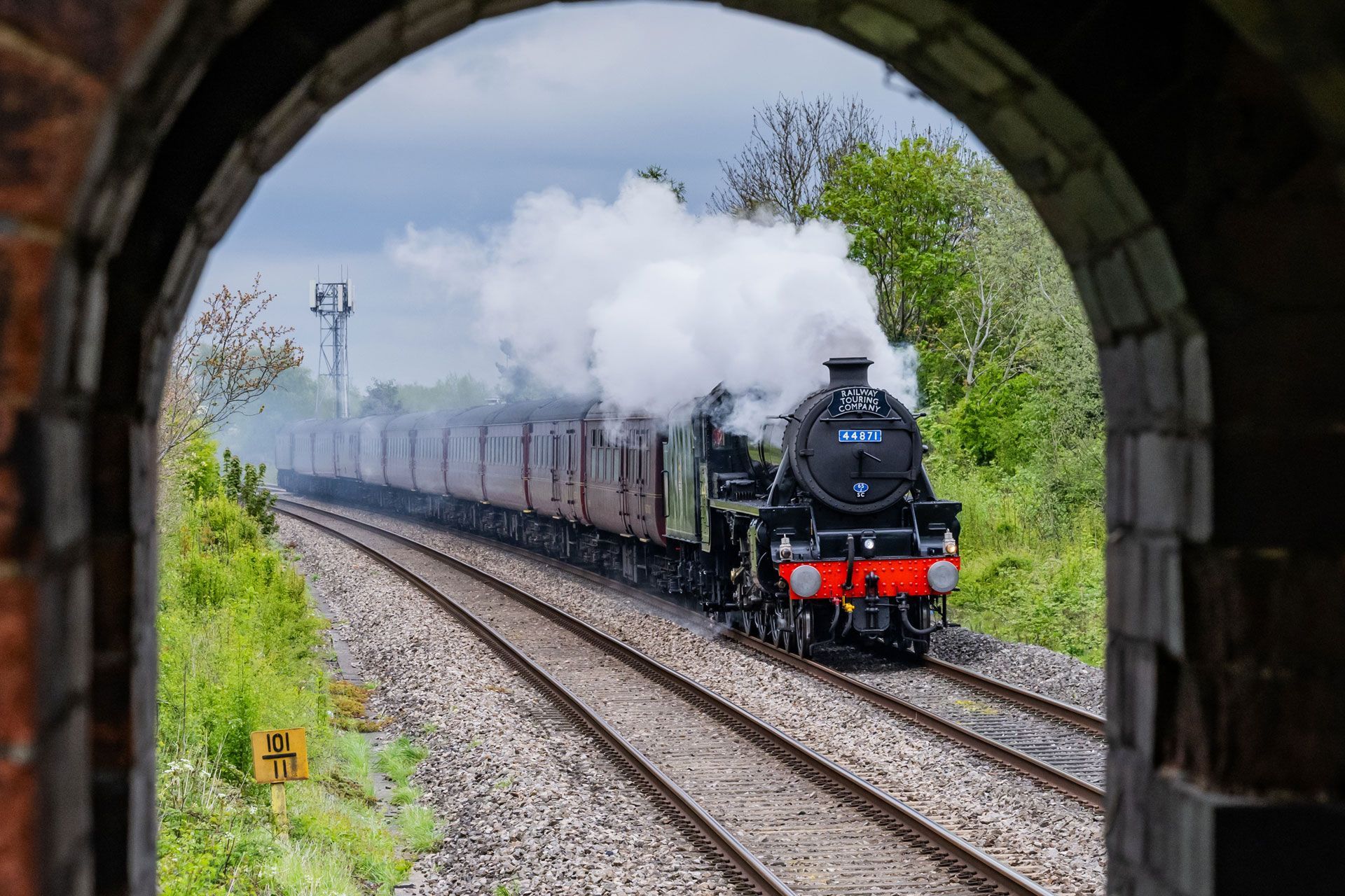 Steam locomotive pulling passenger cars through a tunnel arch, billowing white smoke on curved tracks