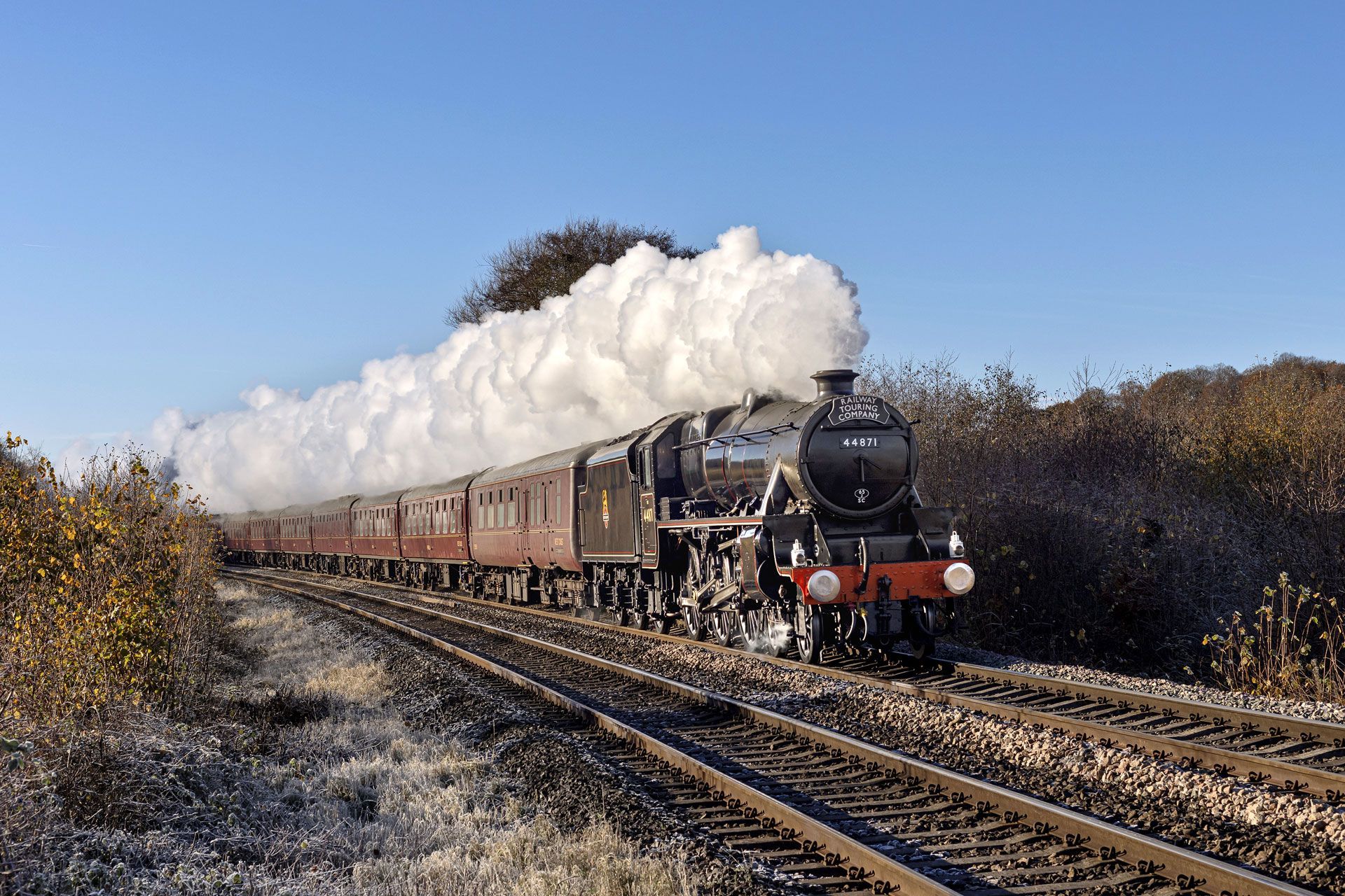 Steam locomotive pulling passenger carriages, billowing white smoke along rural tracks under a clear blue sky