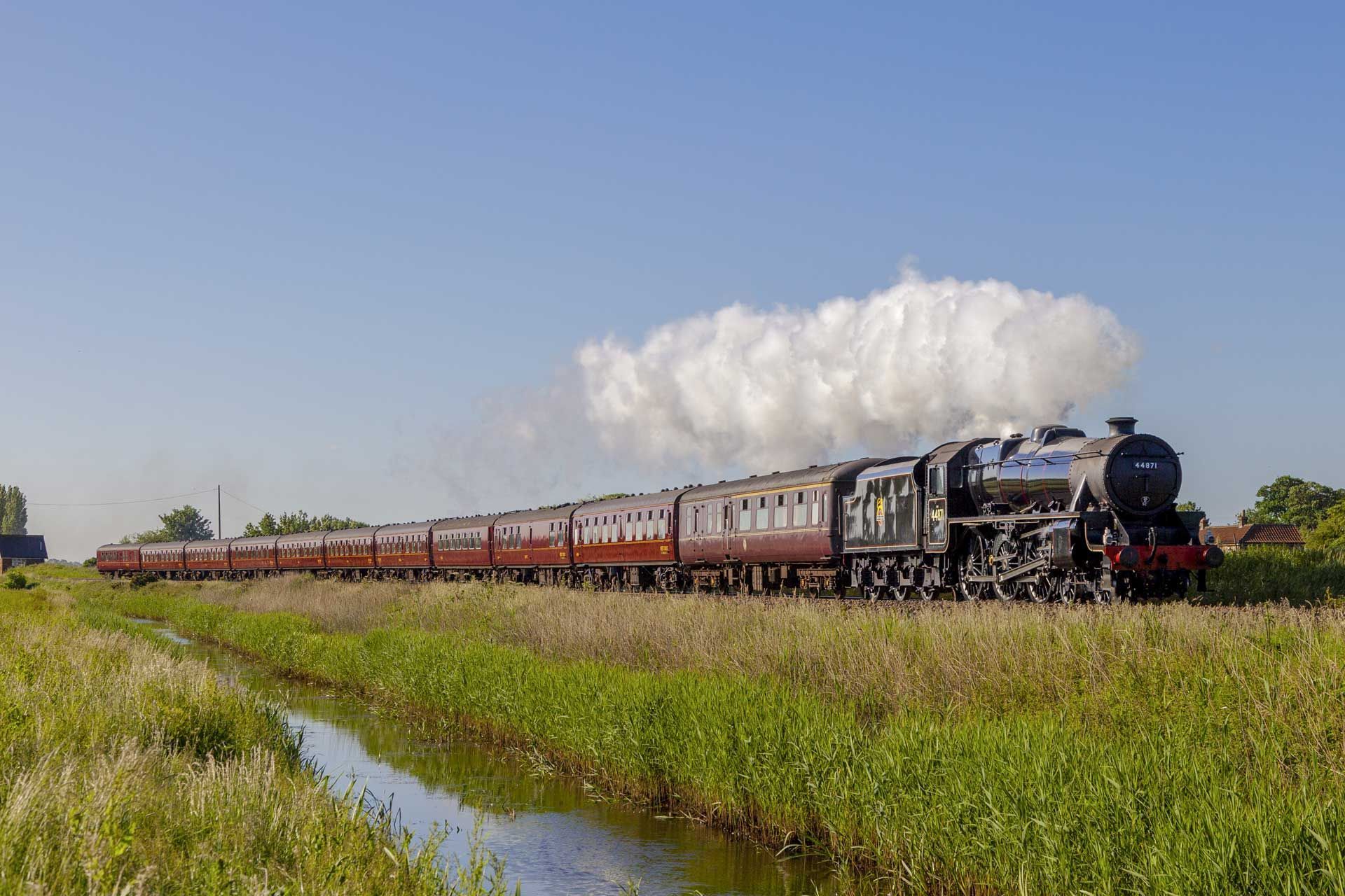 Steam train traveling along a track beside a canal, under a bright blue sky with white cloud.